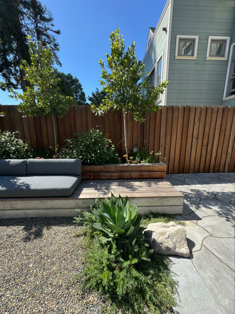 Custom-crafted S4S site furniture beautifully supports cushioned seating and extends linearly, offering a secondary surface for sitting or use as a table. It also transitions seamlessly into the side of a planter that holds trees and florals, while the S4S fencing pattern beyond creates an appealing, patterned backdrop and screen, expertly designed by landscape architect Studio Mala.