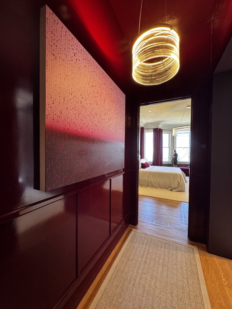 Wood Highlights: The hall of the primary dressing area is intense as it leads to the primary bedroom, by local Castellanos Interiors at the 2026 San Francisco Decorator Showcase. Lacquered paneling with a crisp door casing carries the room's tonal red scheme through the threshold. The effect reads as customization and uniqueness rather than stock materials.